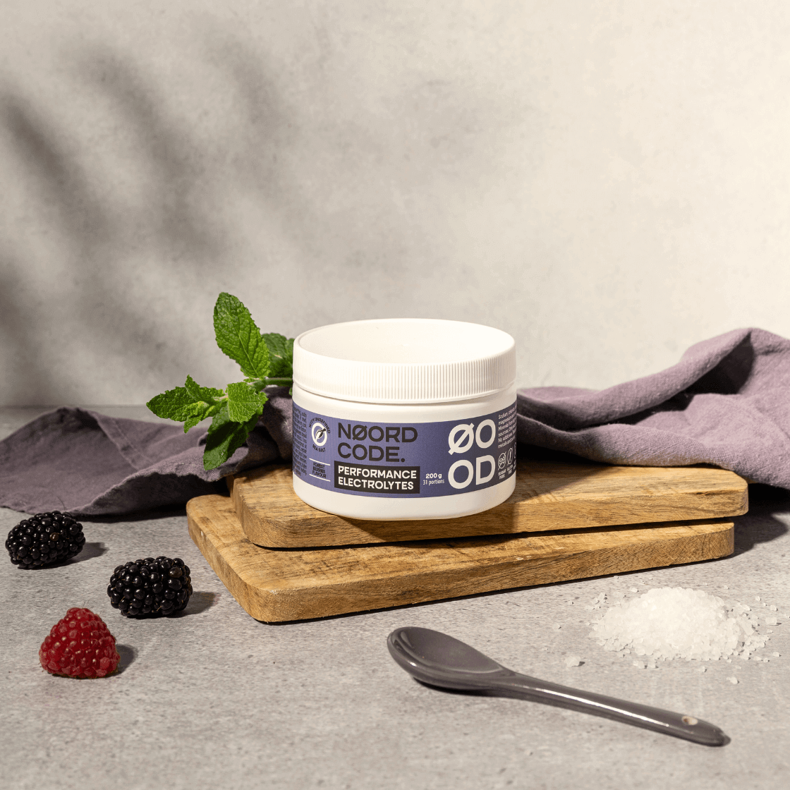 Performance elektrolyty lesné plody na doske, pod nimi lyžička a vľavo černice, maliny a mäta, všetko na neutrálnom pozadí.